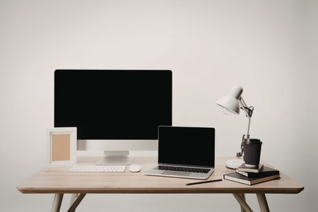 A modern workspace with a computer monitor, laptop, lamp, books, and a mug on a wooden desk against...