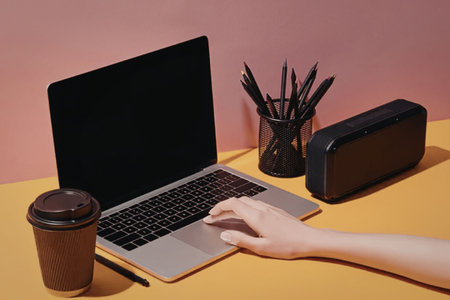 A modern workspace setup with a laptop, hand, coffee cup, speaker, and office supplies on a colorful desk against a pink wall.の写真素材