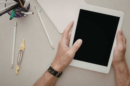 A person is holding a white tablet with a black screen, surrounded by office supplies such as pens,...