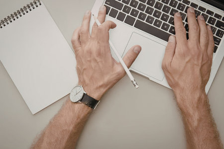 A person working on a laptop with a notepad and pen on the side, typing with one hand and holding a...