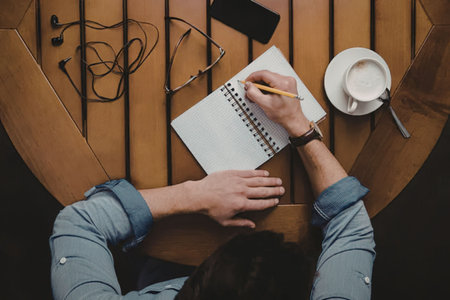 A person sitting at a wooden table, writing in a notebook with a pencil, wearing a blue shirt and a watch, with a cup of coffee and headphones nearby.の写真素材
