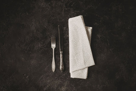 a table setting with a folded napkin and a fork and knife on a dark surfaceの写真素材
