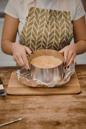 A woman in an apron is preparing a cake in a kitchen, with the cake in a metal pan on a wooden...