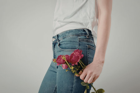 A woman in a white shirt and blue jeans holding pink roses behind her back.