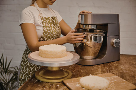 A woman in an apron standing next to a mixer, decorating a cake on a turntable with another cake on...