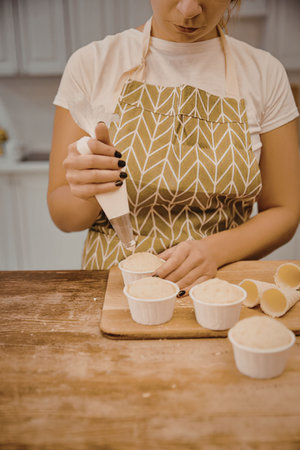 A woman in a green apron is preparing a creamy substance in a kitchen, filling small white cups.の写真素材
