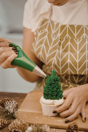 A woman in an apron decorating a cupcake with a green Christmas tree made of frostingの写真素材