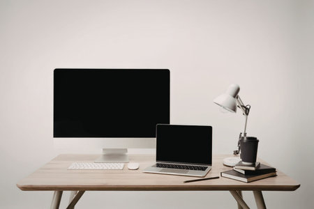 A modern workspace setup with a laptop, monitor, and desk lamp on a wooden desk against a plain...