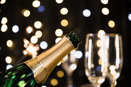a green bottle of champagne with a gold foil top next to two empty wine glasses on a dark background with blurred lights
