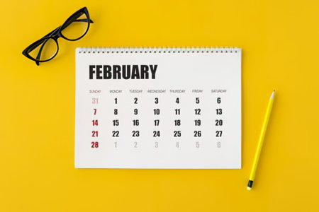 a white calendar page for february is placed on a bright yellow surface alongside a pair of black glasses and a yellow pencil