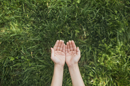 a pair of fair-skinned hands cupped together in an open position, palms up, with fingers slightly spread apart, set against a lush green grassy background with varying shades of green and different textures of blades, suggesting a natural outdoor setting