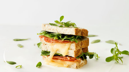 a stacked sandwich made with toasted bread, melted cheese, sliced tomato, and leafy greens, on a white surface with scattered greens
