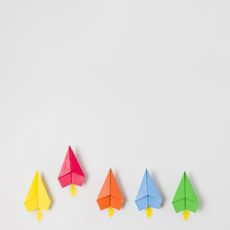 a group of five origami trees in different colors including yellow, red, orange, blue, and green, each with a small yellow trunk, arranged in a row on a plain white surface