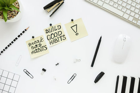 a white desk with office supplies including a keyboard, mouse, pens, pencils, paper clips, and a small plant. There are also two motivational notes that read 'build good habits' and 'break bad habits'.