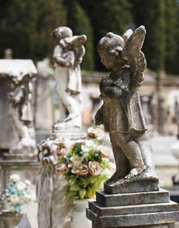 Full view of a carved stone child angel standing on a pedestal in a cemetery with additional child...