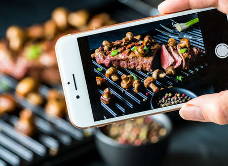 Close up view of a cell phone taking a picture of a strip loin steak on a grill with mushrooms and green onions agains a blurred background of the actual sceneの写真素材