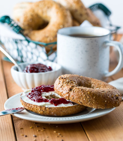 A close up view of a multi grain bagel with cream cheese spread and raspberry jam and surrounded by...