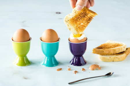 Close up of eggs in egg cups with a hand dipping toast into the yolk of one egg.