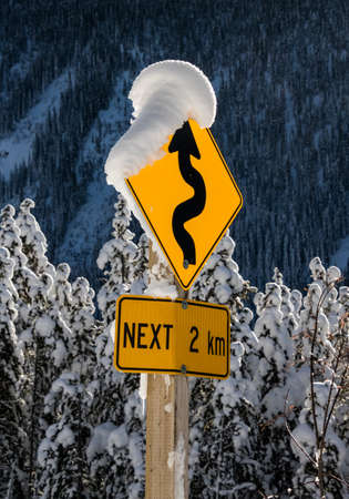 A close up of a road sign that is covered in snow indicating the road is winding for the next two kilometres.の写真素材