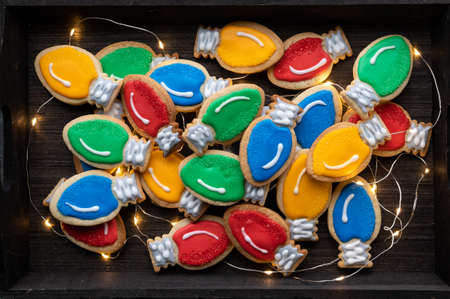 A pile of Christmas lightbulb sugar cookies amongst led string of lights in a dark wooden tray.の写真素材