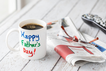 Mug with Happy Fathers Day painted on it filled with coffee, beside a newspaper and remote. Fathers...