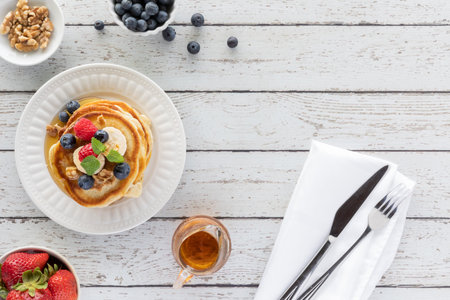 Top down view of a stack of buttermilk pancakes topped with fruit, on a wooden table and cutlery to...