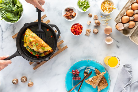 Top down view of hands holding a spatula serving up a freshly made omelette, beside ingredients used...