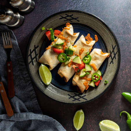 Top down view of a plate of taquitos topped with a cucumber and tomato salsa, served with lime wedges.の写真素材