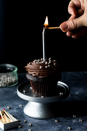 A hand lighting a candle on a chocolate cupcake, against a black background.の写真素材