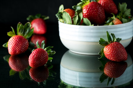 A bowl full of juicy ripe strawberries with some on a glass table.の写真素材