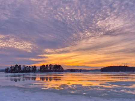 Finnish frozen lake with tree reflections at sunset under dramatic sky.の写真素材