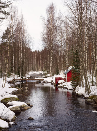 River snowy bank with flowing water and a red cabin and a bridge in viewの写真素材