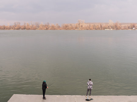 Two figures stand on a concrete platform, fishing in a tranquil river with autumnal trees behind.の写真素材