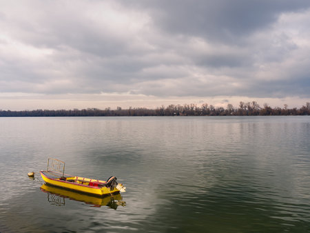 Solitary yellow boat floats on tranquil shore, surrounded by cloudy sky and distant treeline. Zemun, Belgradeの写真素材