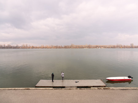 Tranquil Danube river scene with two people on a concrete dock, red boat nearby, overcast sky. Zemun, Belgradeの写真素材