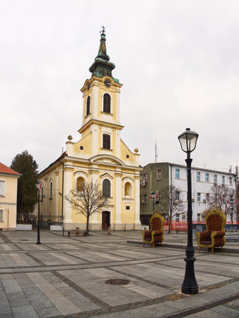 Ornate yellow church with tall spire in town square, surrounded by buildings and street lamps. Zemun, Belgradeの写真素材