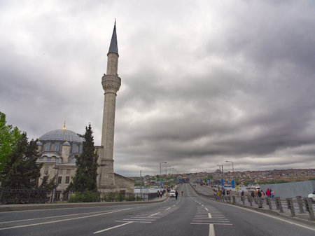 Ottoman-style mosque with minaret along a waterfront road, overlooking a city under dramatic cloudy skiesの写真素材