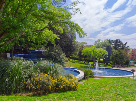 Serene park landscape with a blue circular fountain, lush trees, and colorful flower beds on a sunny dayの写真素材