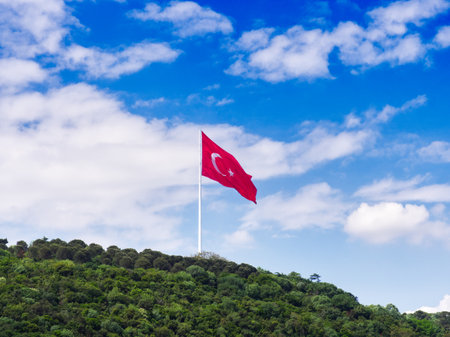 Red Turkish flag with white crescent and star flying on tall pole above lush green hillside under blue skyの写真素材