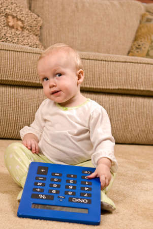 A young baby in a pair of generic PJs sitting on a living room floor with a large oversized calculator. The baby is looking up mischievously.の写真素材
