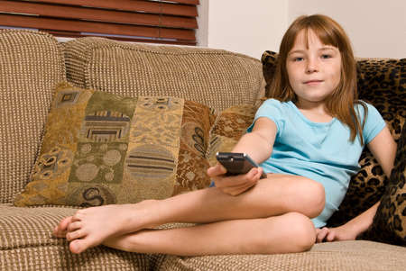A young female child watching television. She is sitting in the corner of the couch holding the T.V. remote control.の写真素材