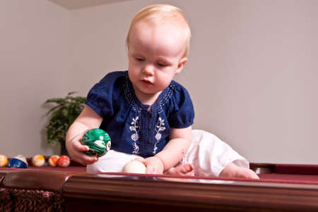 A young child sitting on a billiard table playing with the pool balls. She holds the six ball in here hand and is attempting to drop it in a side pocket of the table.の写真素材