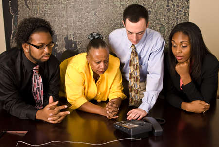 A group of workers stand at a a conference room table during a conference call. All four team members look at the phone with a concerned look on their faces.の写真素材