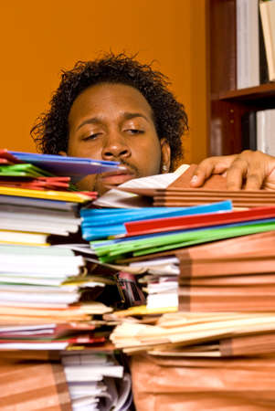 A young African American male buried in work strains to look inside a folder on the top of a stack of colorful folders piled on his desk.の写真素材