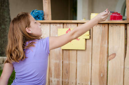 A young girl dipping her paintbrush in red paint as she paints a lemonade stand signの写真素材