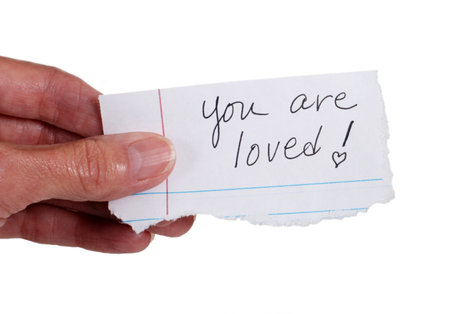 Close up of a hand holding a note with the words: 'You Are Loved'.  Isolated on white.