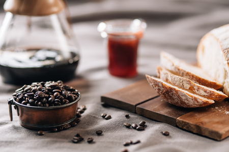 A tabletop scene with coffee beans in a pot, some bread, jam and coffee on a table clothの写真素材