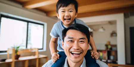 Jubilant Asian father having a blast with his young son in the living room, carrying him on his back and enjoying quality time at home.の素材