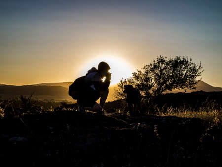 Photographer silhouette on the mountain is taking photos to a big dog, shooting at the landscape with sunset or sunrise. Photography concept. Sierra de la ventana, Buenos Aires, Argentina.の写真素材