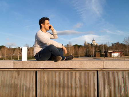 Young handsome caucasian man speaking on his phone, seated at Madrid Rio with the Almudena Cathedral...